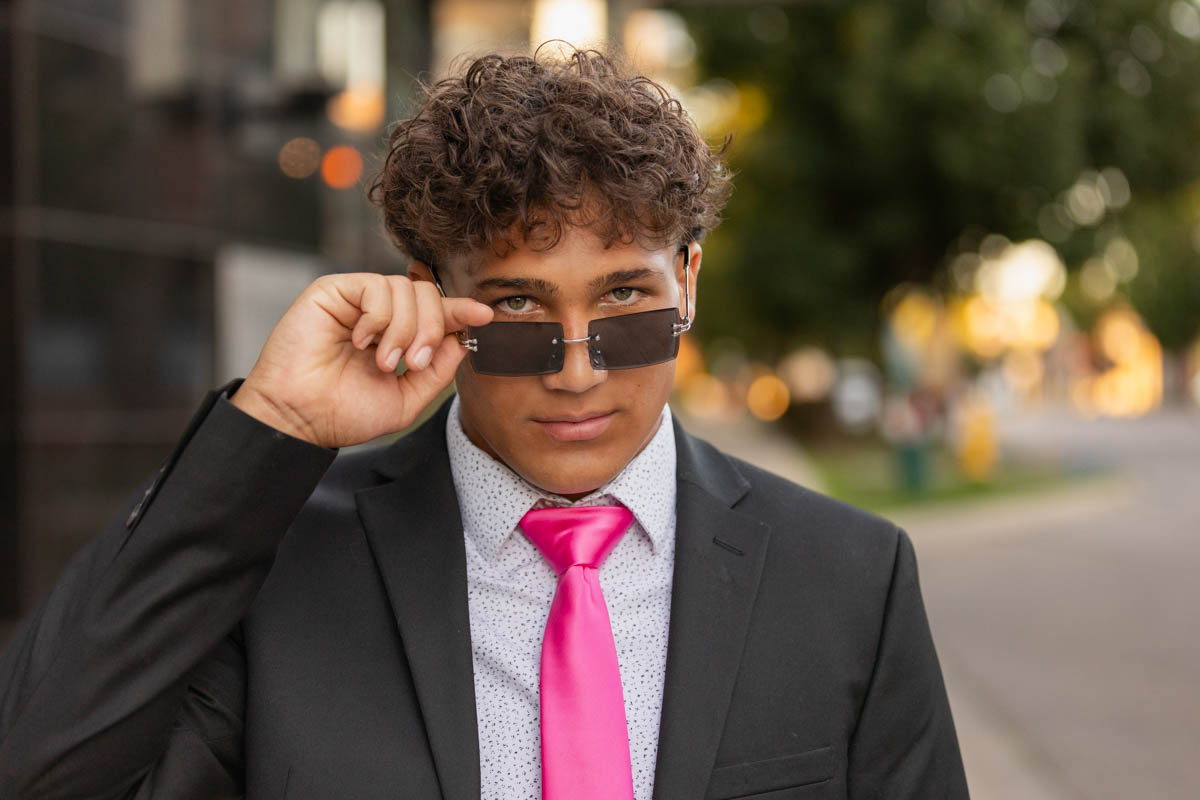 High school senior boy in a suit with a pink tie, lifting sunglasses and looking confidently at the camera.