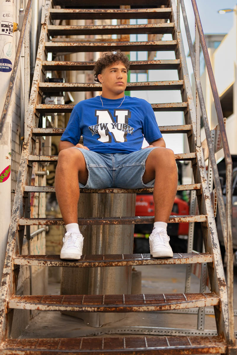 High school senior boy in a blue New York t-shirt and jean shorts sitting on a rusty metal staircase.