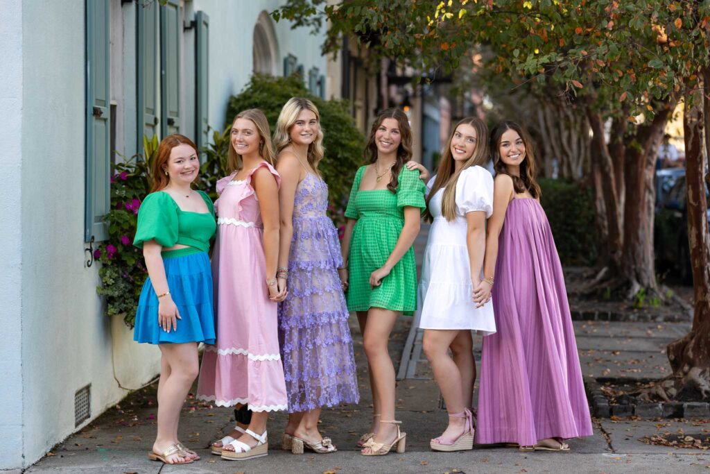 A group of six high school senior girls from the MBP Senior Rep Team posing together on a charming city sidewalk, dressed in colorful and stylish dresses. They are smiling, holding hands, and enjoying their senior photography session with Megan Bowers Photography.