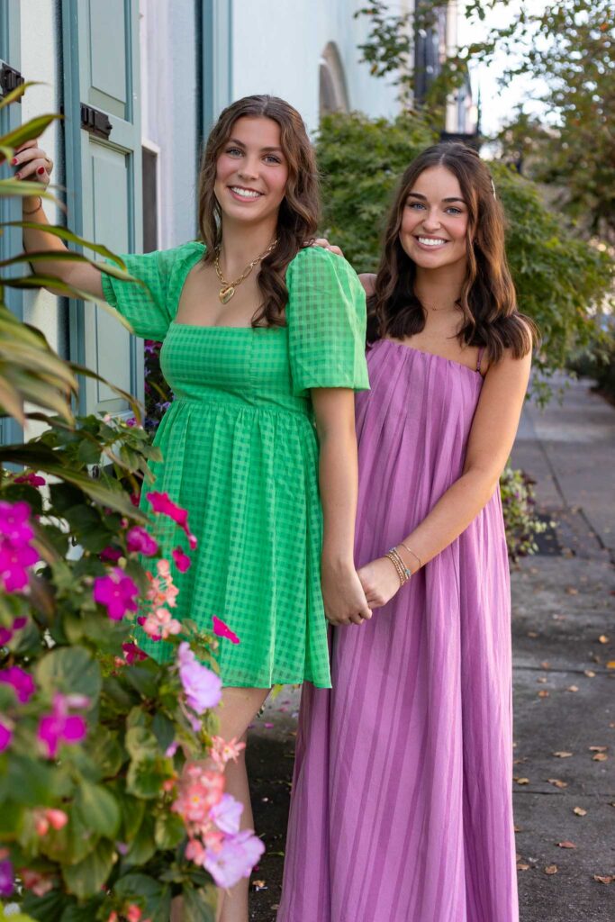 Two high school senior girls from the MBP Senior Rep Team smiling and holding hands during a stylish senior photography session. One wears a green gingham dress, and the other a flowing purple dress, posing by a charming city sidewalk with flowers and pastel-colored buildings in the background.