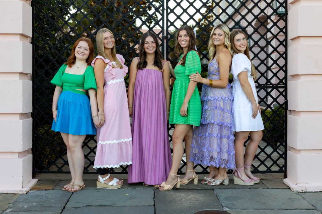 A group of six high school senior girls from the MBP Senior Rep Team posing together in stylish, colorful dresses in front of an elegant black iron gate. The team smiles confidently, celebrating their senior year with a professional photoshoot.