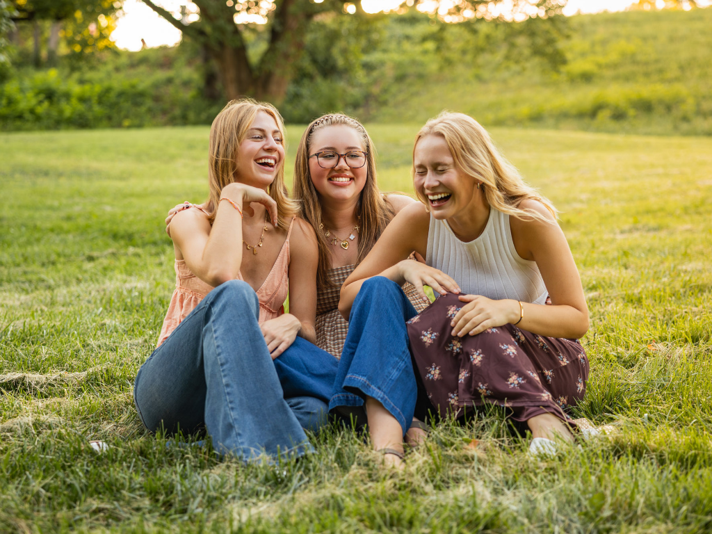 Three senior girls sit close together in the grass, laughing during a summer group photoshoot.