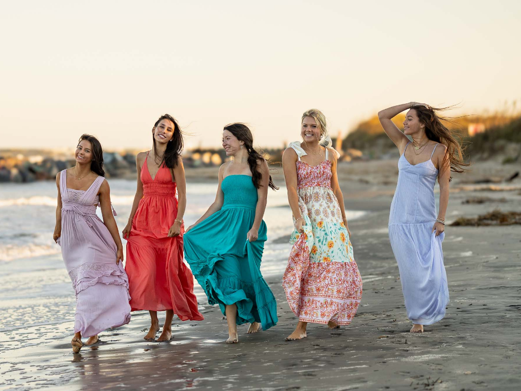 Five senior girls walk barefoot on the beach at sunset in flowing maxi dresses during a styled group photoshoot.