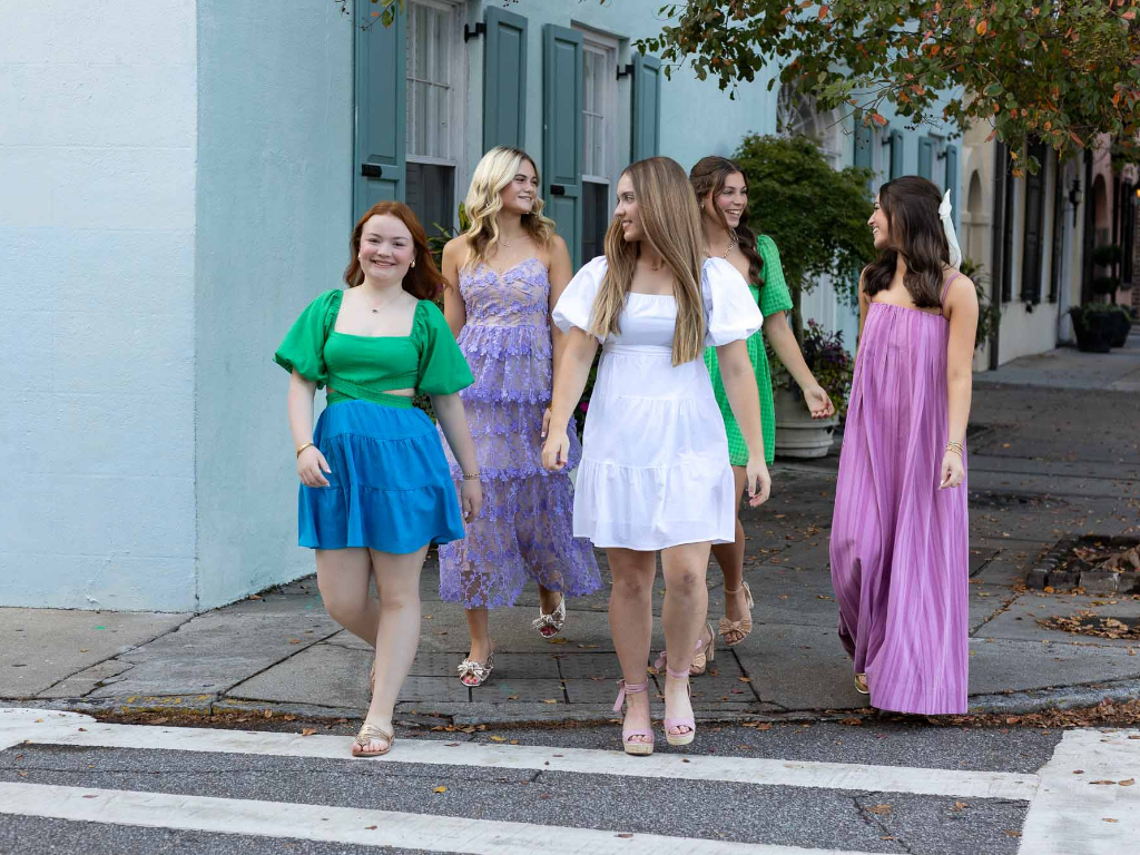 A group of senior girls laugh and talk as they cross the street together in bright spring dresses.