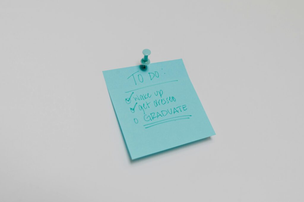 A blue sticky note pinned to a white surface with a handwritten graduation to-do list that includes ‘Wake up,’ ‘Get dressed,’ and ‘Graduate.’ A fun and motivational reminder for high school seniors preparing for graduation day. Perfect for Graduation Etiquette Guide, senior photography, and celebration planning.