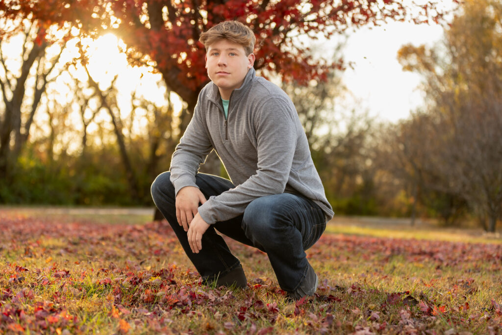 Senior boy in gray sweater kneeling in autumn leaves for outdoor shoot