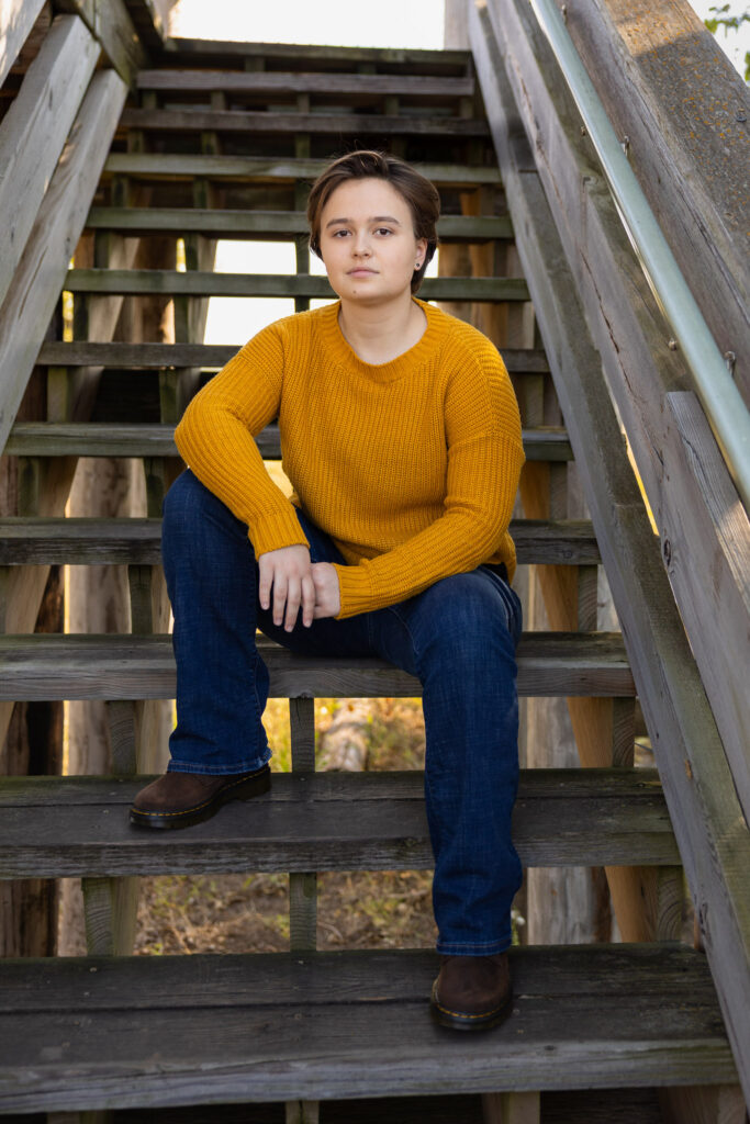 Amelia wearing a mustard sweater sitting on wooden stairs during senior portraits in Lawrence, KS