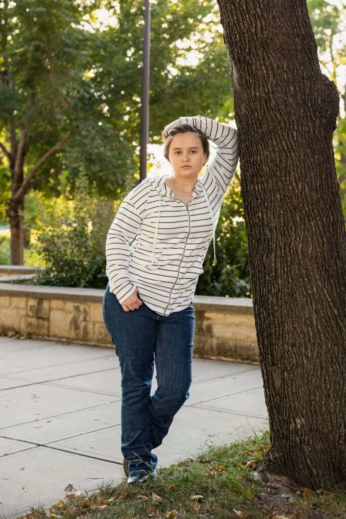Amelia posing by a tree in a striped hoodie during senior portraits on the University of Kansas campus