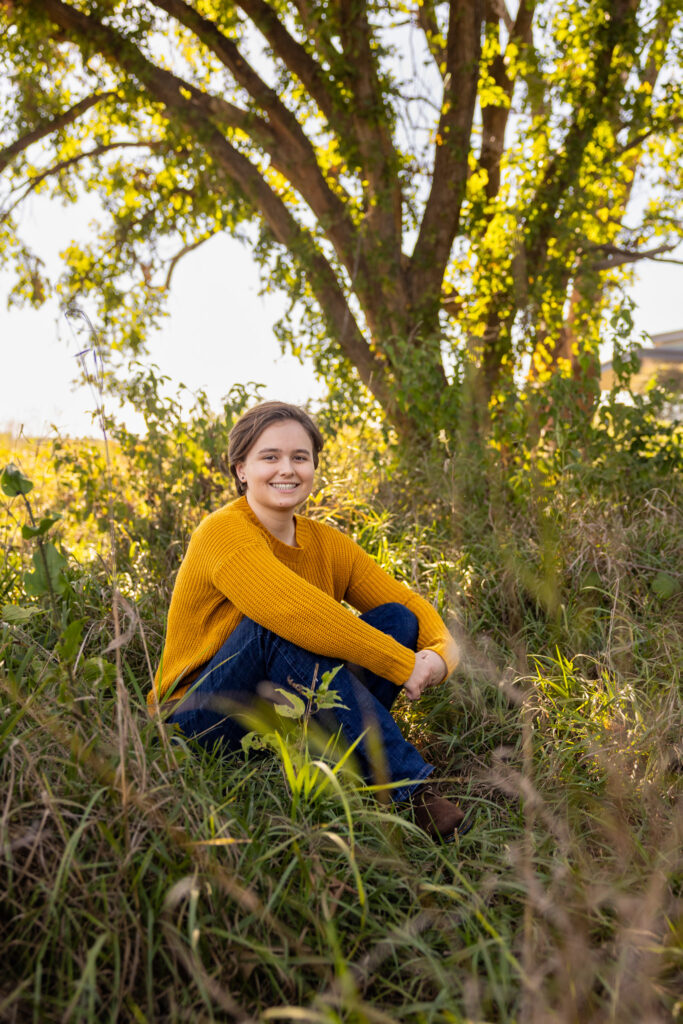Amelia sitting in tall grass under a tree during golden hour senior photos in Lawrence, KS