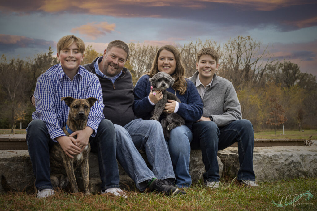 Family of four sitting on a bench outdoors with two small dogs during a senior family photo session in Lawrence KS
