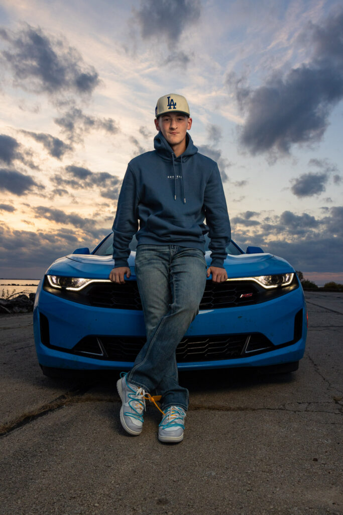 High school senior boy in an LA cap and navy hoodie sitting on the hood of a blue Chevrolet Camaro at sunset for his Lawrence, KS senior portrait session