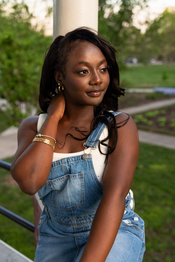 High school senior girl in denim overalls and gold jewelry posing outdoors at golden hour for her Lawrence, KS senior portrait session