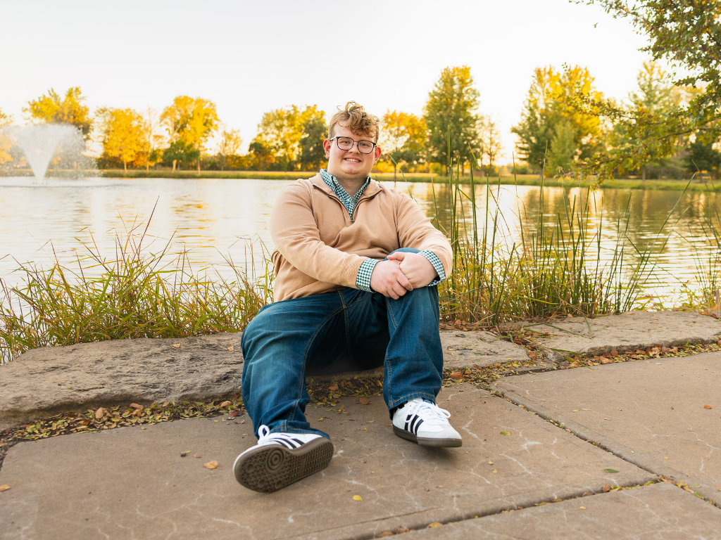 Senior in a tan quarter-zip sitting by a pond with a fountain and golden fall trees at the Lawrence Arboretum