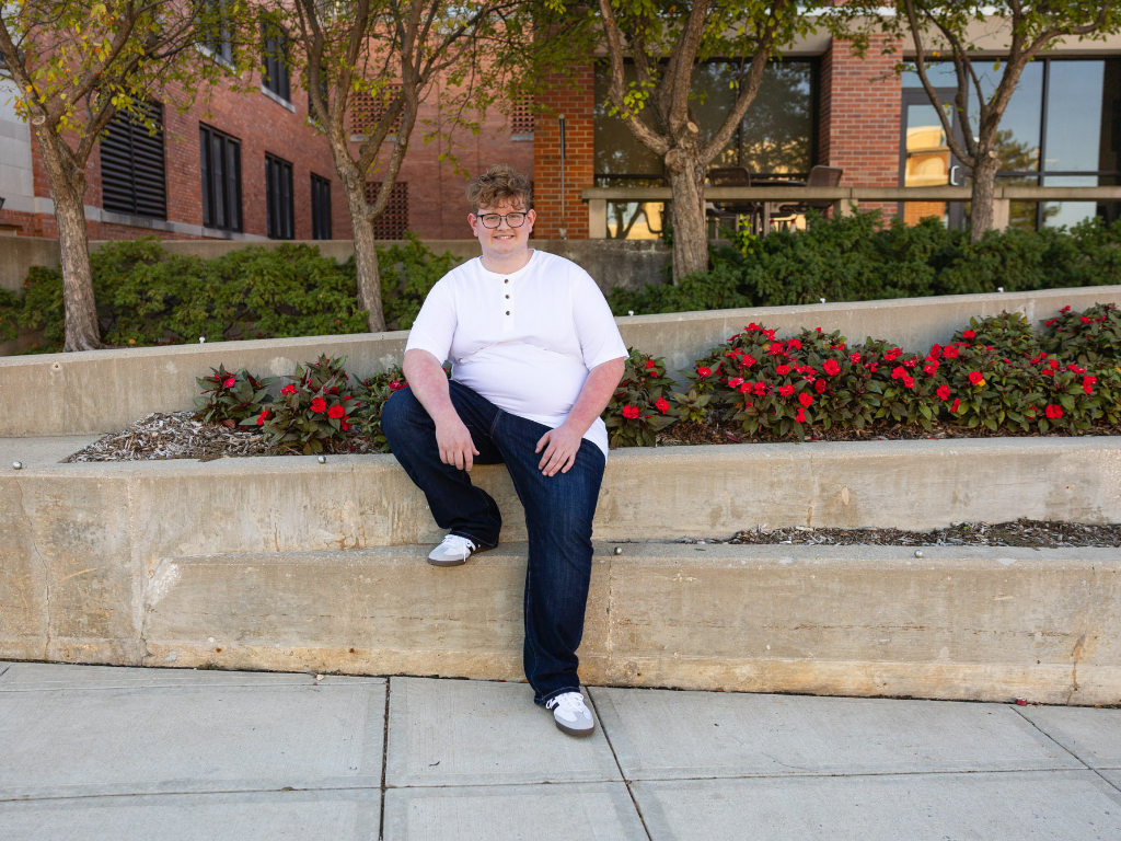 Senior guy in a white henley sitting on a concrete planter with red flowers outside a brick building at the University of Kansas