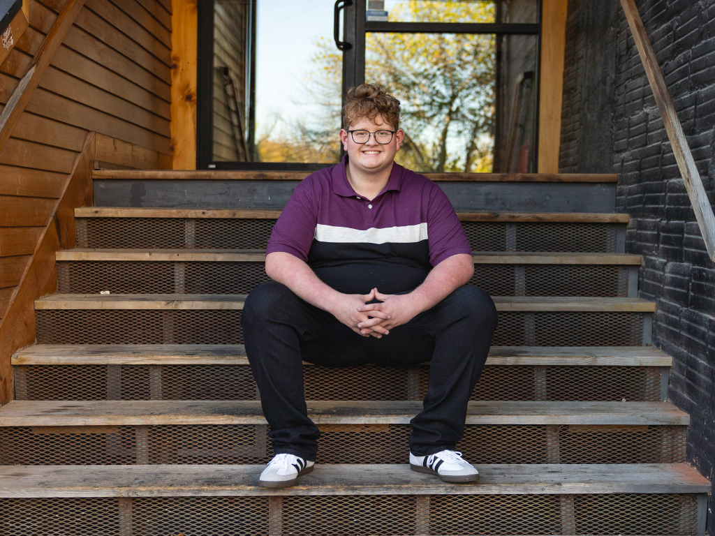 Senior in a purple polo sitting on metal steps outside a brick building for his senior portraits in Lawrence, Kansas