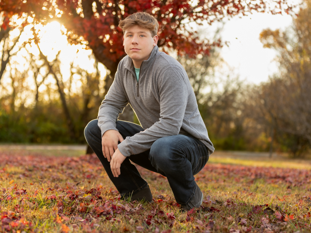 High school senior in a gray quarter-zip and dark jeans kneeling among red fall leaves at golden hour for his Lawrence KS senior pictures.