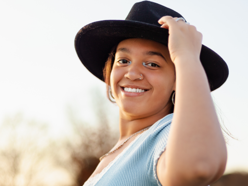 Smiling senior girl in a wide-brim black felt hat and light blue ribbed top holding the brim during her Lawrence senior session at golden hour.