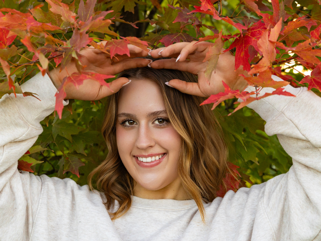Smiling high school senior girl in a cream sweatshirt holding red and orange fall leaves above her head during her Lawrence senior session.