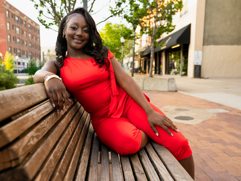 Tay during her Lawrence senior session sitting on a wooden bench along Mass Street, downtown storefronts behind her, in a red off-the-shoulder jumpsuit.
