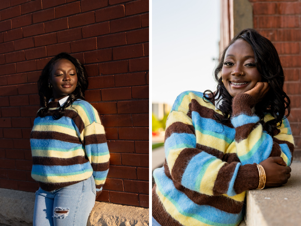 Tay leaning against a dark red brick wall in a blue, brown, and yellow striped sweater and ripped jeans, smiling with her hand under her chin.