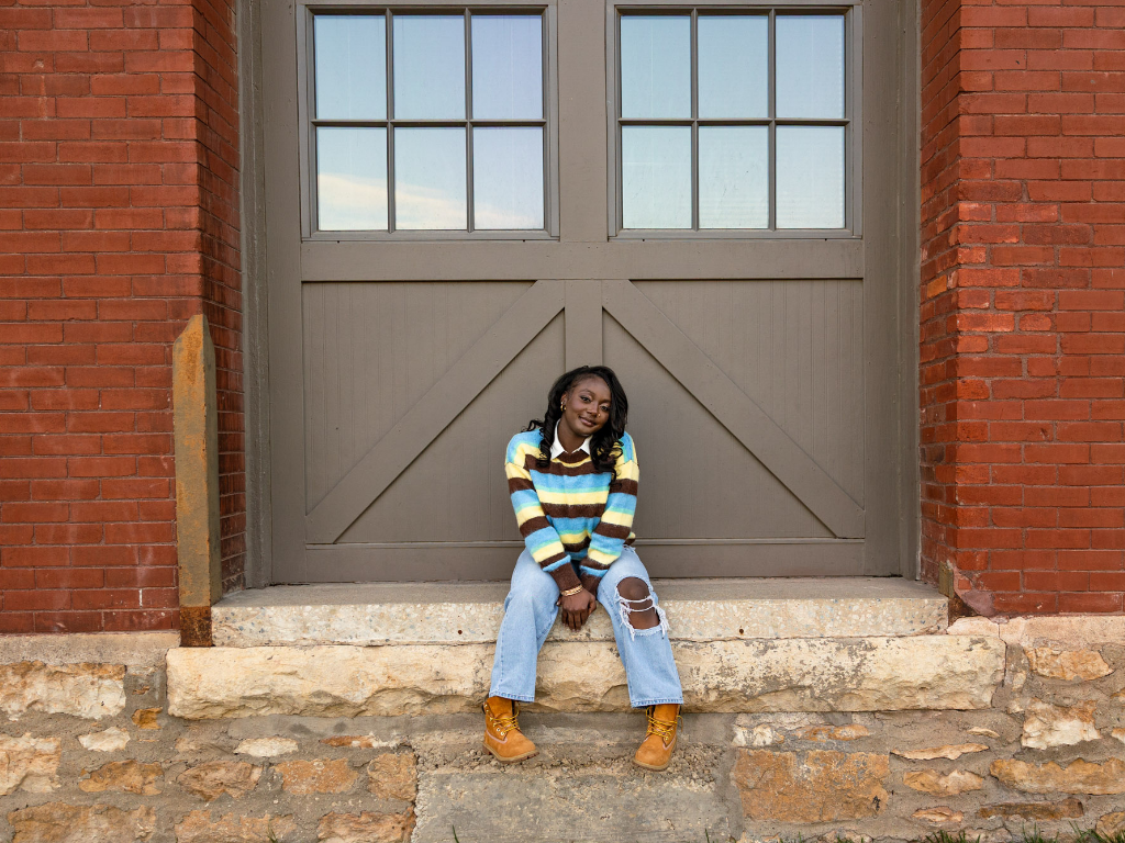 Tay sitting on a stone ledge in front of a brick building in a striped knit sweater, ripped jeans, and tan boots.