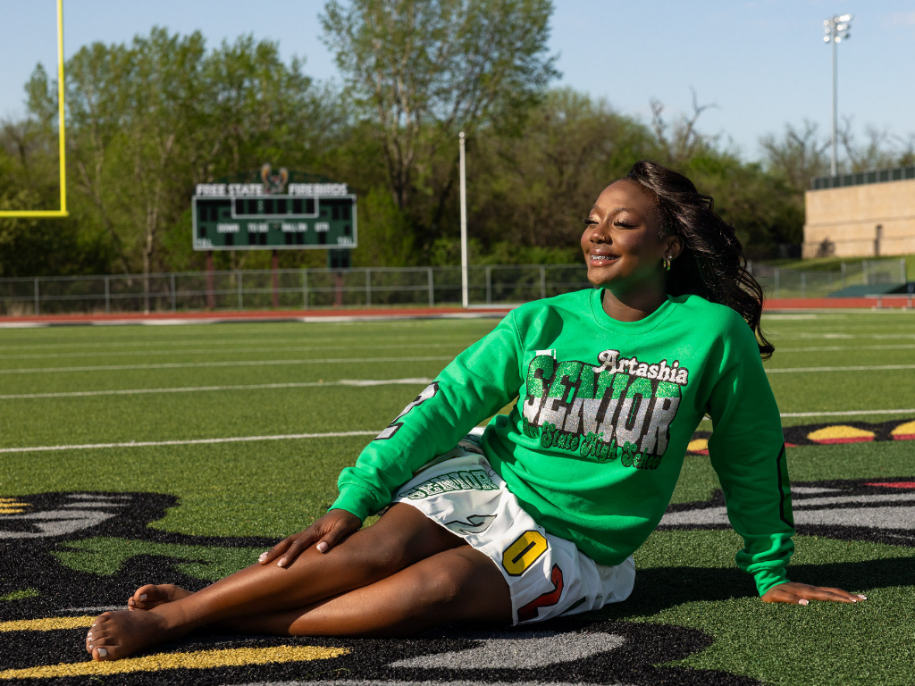 Free State senior Tay during her spring senior session in Lawrence, Kansas, sitting on the football field in a green custom senior sweatshirt and white 2026 skirt.