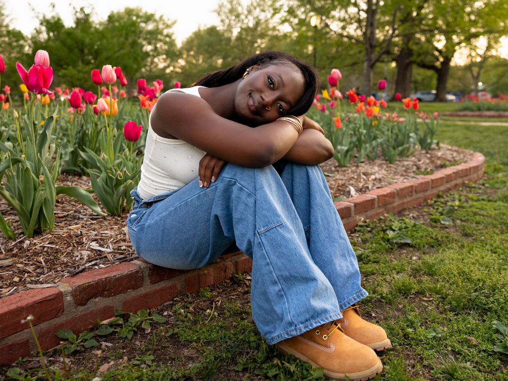 Tay resting her head on her knees beside a row of pink and red tulips at South Park in Lawrence, in a white tank and light-wash denim.