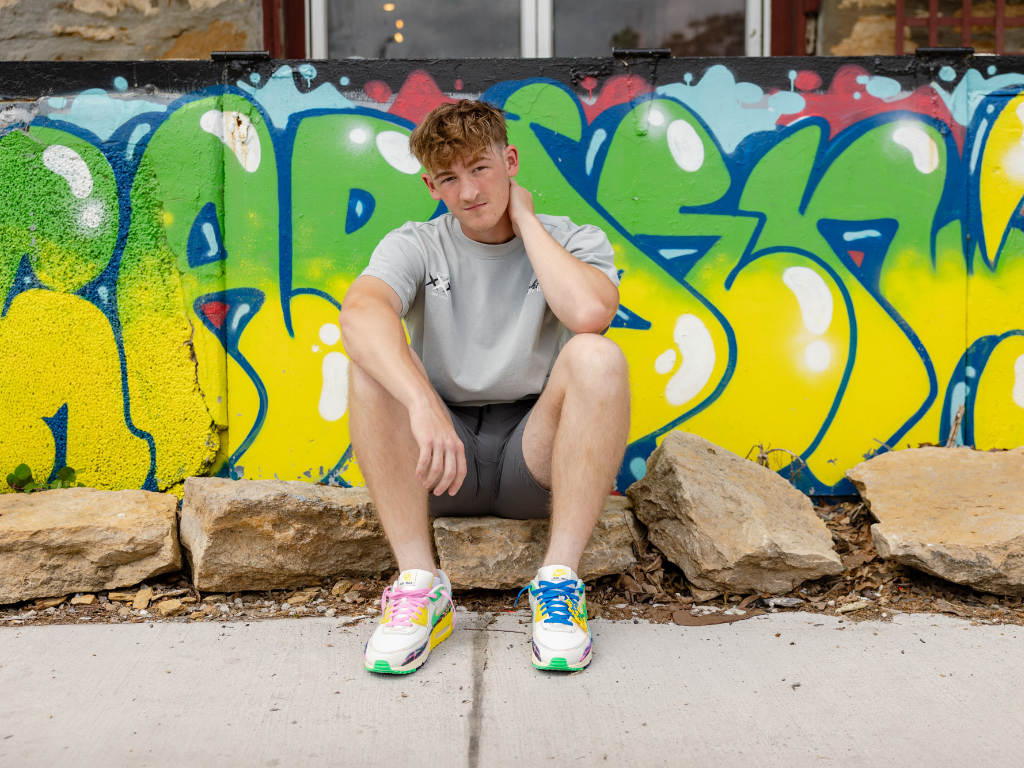 Senior guy sitting on a stone ledge in front of a bright graffiti wall, wearing colorful sneakers.