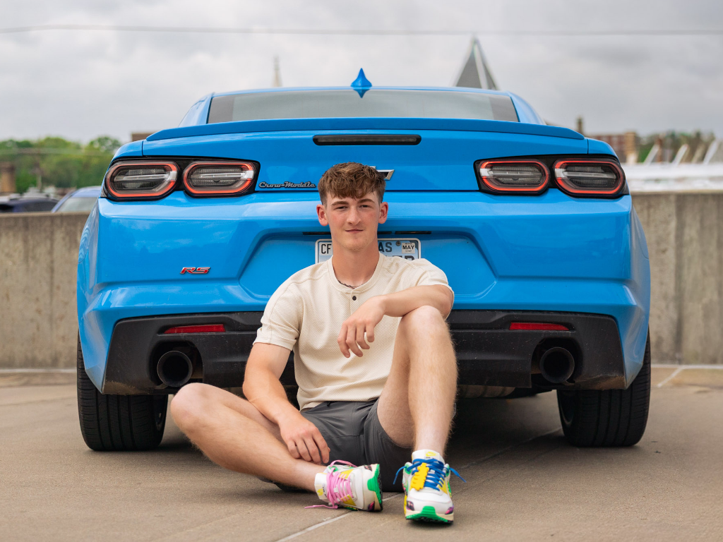 Senior boy sitting cross-legged behind a blue Camaro in colorful sneakers, rooftop parking setting.