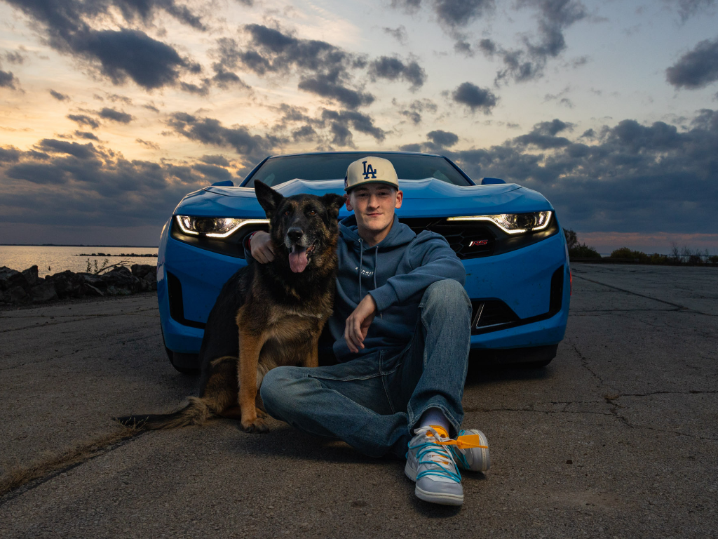 Senior guy in a ball cap hugging his German shepherd in front of his blue car by the lake at sunset, Lawrence KS senior pictures