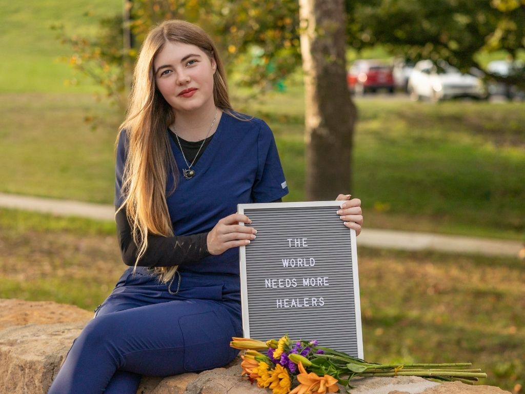 Lily in navy scrubs sitting on a stone wall, holding a letter board that reads “The world needs more healers”